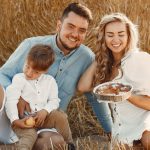 Smiling family of three enjoying a sunny day picnic with pastries on a hay field.